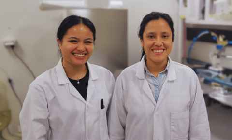 Two smiling women in a lab, wearing white lab coats.