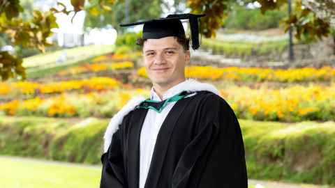 Head and shoulders image of graduate Isaac Hunter in academic regalia against a green and flowery park background.