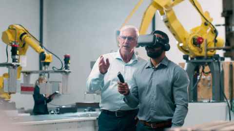 Two men have a discussion in a factory with yellow robot machinery behind; one wears VR goggles