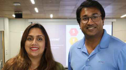 A smiling couple in a classroom with a slide on a screen behind them - the word 'STEM' can be seen.