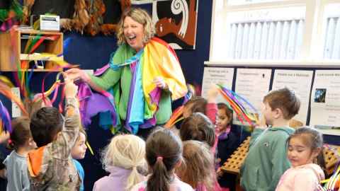 A teacher in bright, colourful cloaks laughs and in a lively way in front of a group of animated children in a classroom.