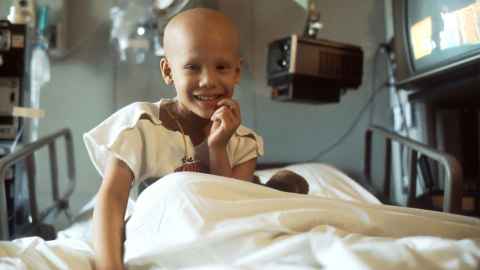 Child with no hair sits on a hospital bed, smiling