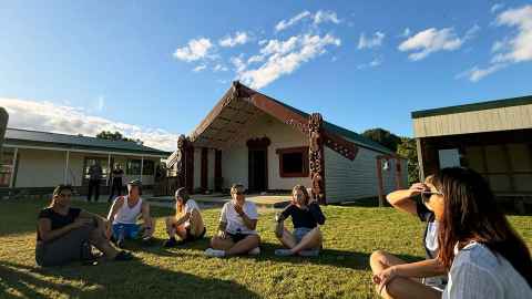 Students sit in a circle on the grass in front of the wharenui at ōwhakatoro Marae, Rūātoki.