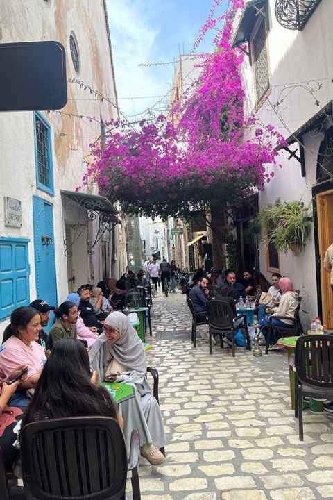 A cobbled street with people sitting outside at cafe tables, and scarlet bougainvillea behind.