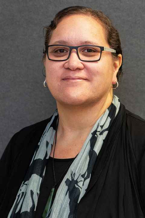 Head and shoulders of Professor Krushil Watene wearing a black top, grey and black scarf and black glasses.