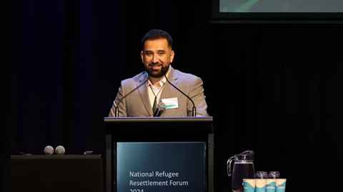 Rabia Talal Almbaid standing at a podium wearing a brown suit, white shirt and pounamu necklace.
