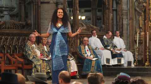 Selina Tusitala Marsh wearing a long blue dress and chiffon wrap, with a large shell necklace, performing a poem at Westminster Abbey in front of the late Queen Elizabeth and other dignitaries and clergy.
