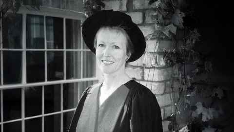 Vivienne Gray wearing academic regalia and standing in front of a window and brick wall.