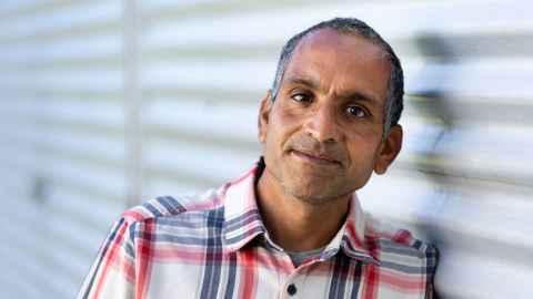 A portrait shot of Dr Ritesh Shah wearing a red, blue and white checked shirt and leaning against blurred white blinds.