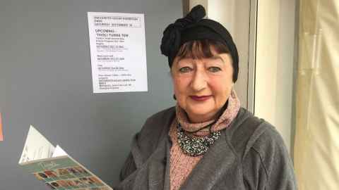 A head and shoulders of Liz Eastmond wearing a rose pink knitted jersey, greeny jacket, chunky necklace and with her hair in a black turban.