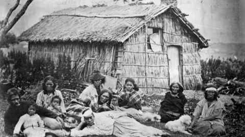 A black and white historical photo of Māori whānau outside a raupō house. Photo: Alexander Turnbull Library, Mrs Scott Collection.