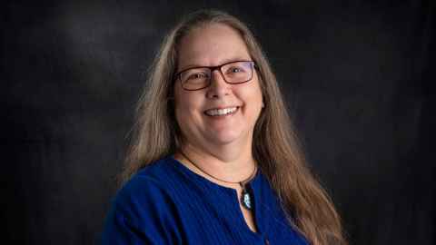 Head and shoulders of Dr Deborah Elms wearing a blue, long-sleeved top and a pendant. Dr Elms has long brown hair and glasses.