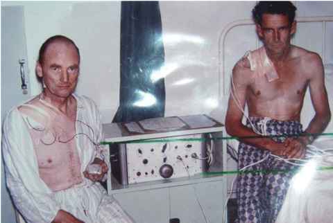 Two guant looking patients sit on beds in pyjamas with a large external pacemaker between them. Historical photo