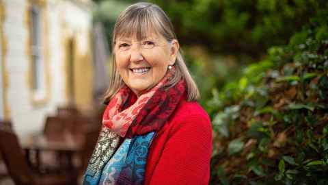 Professor Carol Mutch in the grounds of Old Government House, wearing a red cardigan and a colourful scarf.