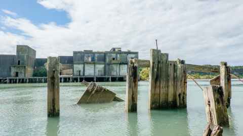 In the background is an old freezing works building, across the Patea River. In the foreground broken wooden pilings emerge from the water. 