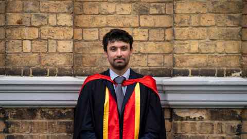 Mathias wearing graduation robes in front of a yellow brick wall