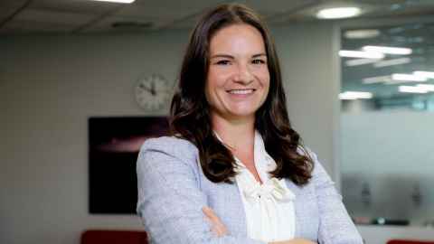 A head shot of Kelly Barton. She has shoulder-length dark hair and wears a pale jacket and white shirt.. She is standing in front of a wall that has a clock on it.