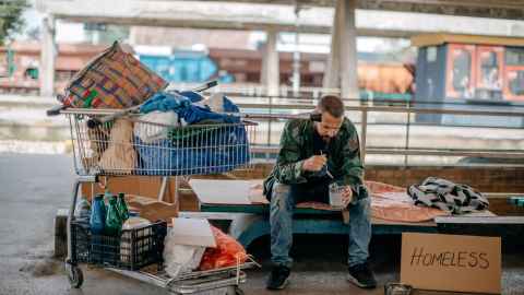 Man on street bench, with belongings in shopping trolley, and homeless sign