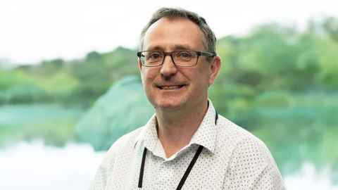Portrait of Associate Professor Barry Milne wearing glasses, a white open-collared shirt and a lanyard round his neck against a green, water and bush background.