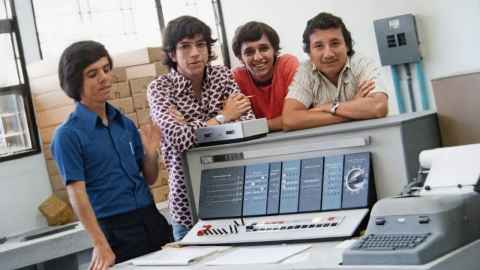 Four young men stand around an old fashioned looking computer