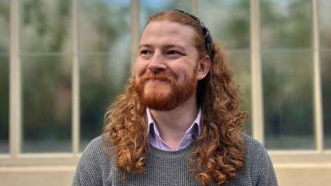 PhD student Nicholas Ringwood with long, ringleted red hair,  a red beard, a lilac collared shirt and a grey jersey.