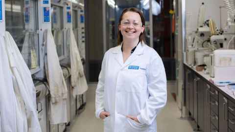 Associate Professor Erin Leitao. Pictured in the foreground of a laboratory. Wearing a white lab coat. Other lab coats are hanging to one side of her. On the other side is a bench with lab equipment.