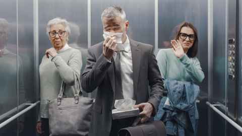 Three people in an elevator; one sneezing, two others pulling away. 