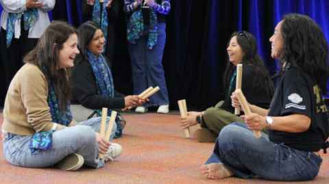 Image of people sitting on the ground playing tī rākau (Māori stick game)