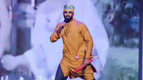 Nimish Milan Singh performing a rap in an orange traditional tunic on stage with a microphone.