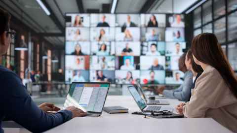 Group of people at business table, formally dressed, facing another group of people on large screen. 
