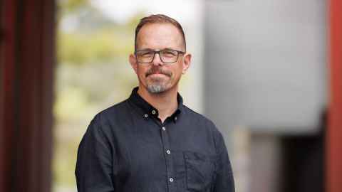 Head and shoulders portrait of Professor Luke Goode wearing a black, collared, long-sleeved shirt against a blurred, outdoor background.