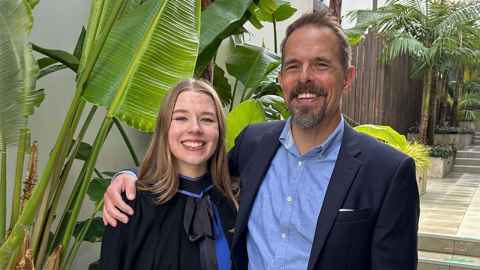 Luke Goode, wearing a blue shirt and black jacket, and his daughter Charia in graduation cap and gown with a tropical garden background.