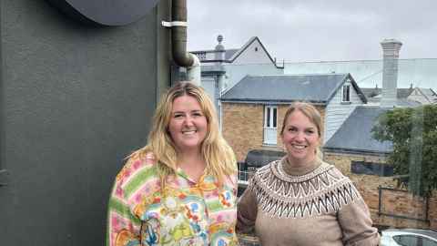 Photo of two women smiling at the camera. They are standing outside and in the background there are residential properties. 
