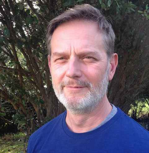 A head and shoulders of Professor Neal Curtis wearing a blue t-shirt against a green background.