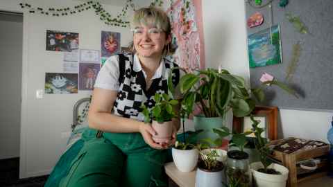 Person sits on their bed leaning over their desk slightly with plants all around them. They are smiling at the camera.