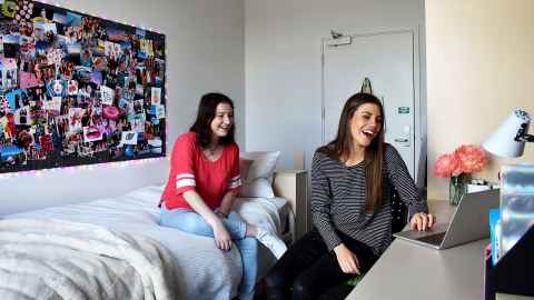 Two residents in a room. One is sitting at the desk looking at a computer and laughing while the other sits on the bed also laughing towards the computer. 