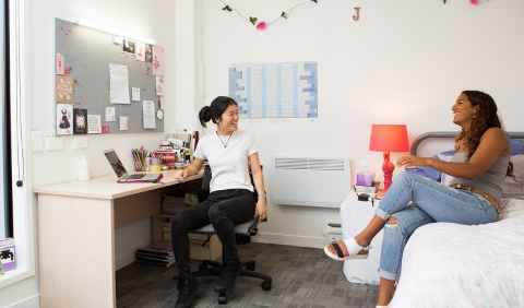 Two students in a bedroom. One is sitting cross legged on a bed and the other is seated in a desk chair.