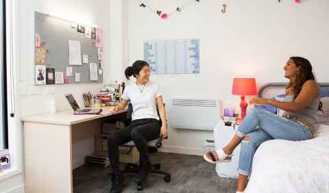 Two students in a bedroom. One is sitting cross legged on a bed and the other is seated in a desk chair. 