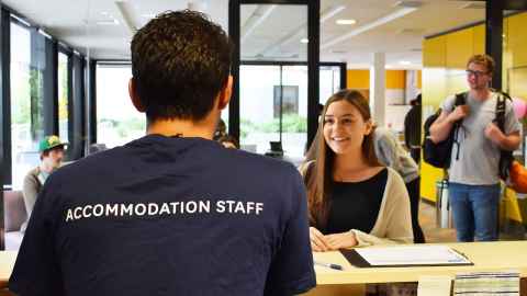 In the foreground a person with their back to camera is standing behind a reception desk and speaking to someone on the other side. Their shirt reads "Accommodation Staff". Other people appear to be walking through the lobby area.