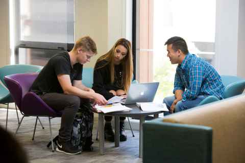 Three students sitting huddled around a laptop. 