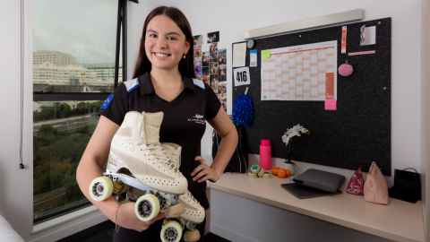 Accommodation resident in her room in front of her desk. She is holding a pair of rollerskates and wearing her NZ competition jersey. 