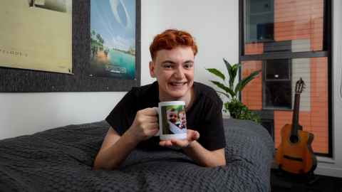Joel lays across his bed smiling and holding a mug with a family photo on it. 