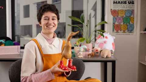 Noa is sitting cross legged in a chair while wearing mustard yellow overalls and a white button up. They hold a wood goose with polka dot. Text reads "Bring your silly creatures. We'll sort the rest" red boots