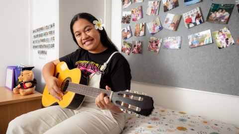 Sala sits on her bed while holding a guitar across her lap.