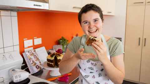 Rose is in her kitchen wearing a green shirt and apron. She is holding a slice of cake, about to eat it.