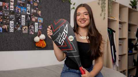 Saffiya is sitting on her bed wearing her taonga, a black top and holding her waka paddle.