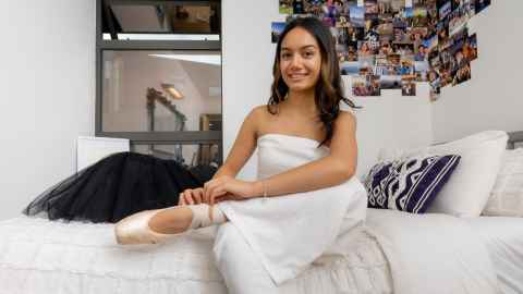 Brianna sits on her bed with one leg crossed over the other and puts on her pointe shoes