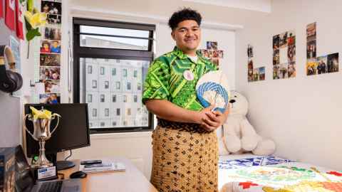 Lolo stands in his room, holding a fan and wearing a ta'ovala