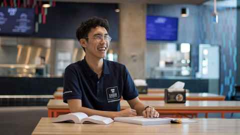 Resident Adviser is in the Waipārūrū dining hall. In their RA uniform and with books in front of them, they are looking in the distance with a big smile. 