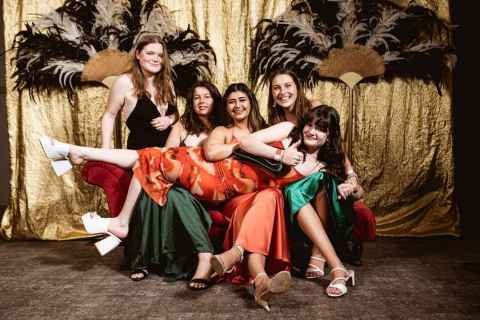 Five students pose in front a golden curtain and feather fans. They are wearing ball gowns. Four of them are sitting on a red bench while one is laying over the laps of the seated people. 